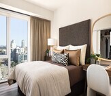 Chic and cozy bedroom featuring a plush bed with decorative pillows, elegant desk with mirror, and a stunning city view through large windows. - Ancora